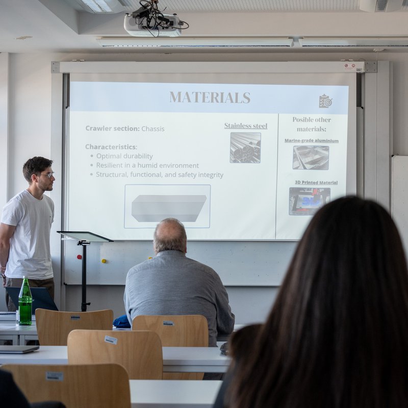 Ein Raum mit Studierenden, vorne an der Wand stehen Studierende und halten einen Vortrag