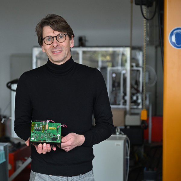 Der Mann in schwarzem Rollkragenpullover hält eine elektronische Platine in den Händen und lächelt freundlich.