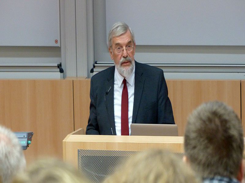 Ein Mann mit roter Krawatte steht am Rednerpult eines Hörsaals und blickt auf seine Zuhörenden.