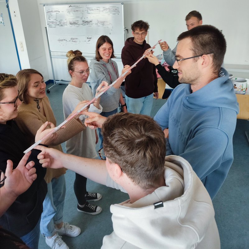 acht Personen halten einen Zollstock gemeinschaftlich hoch als eine Aufgabe zum Teambuilding