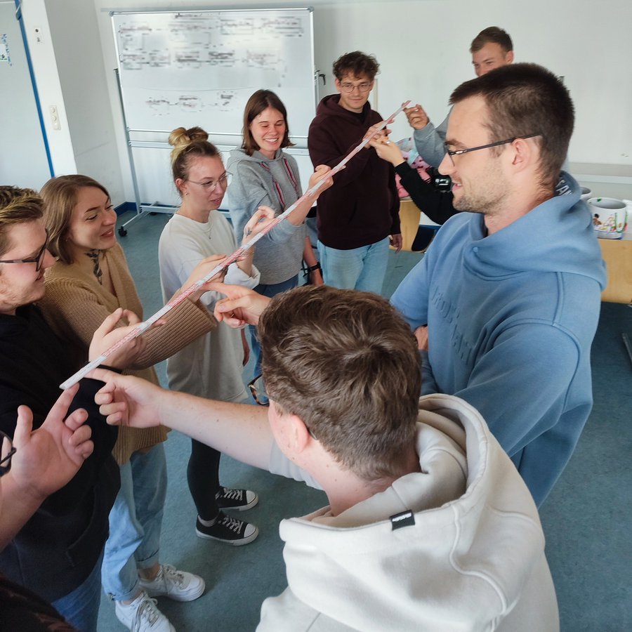 acht Personen halten einen Zollstock gemeinschaftlich hoch als eine Aufgabe zum Teambuilding