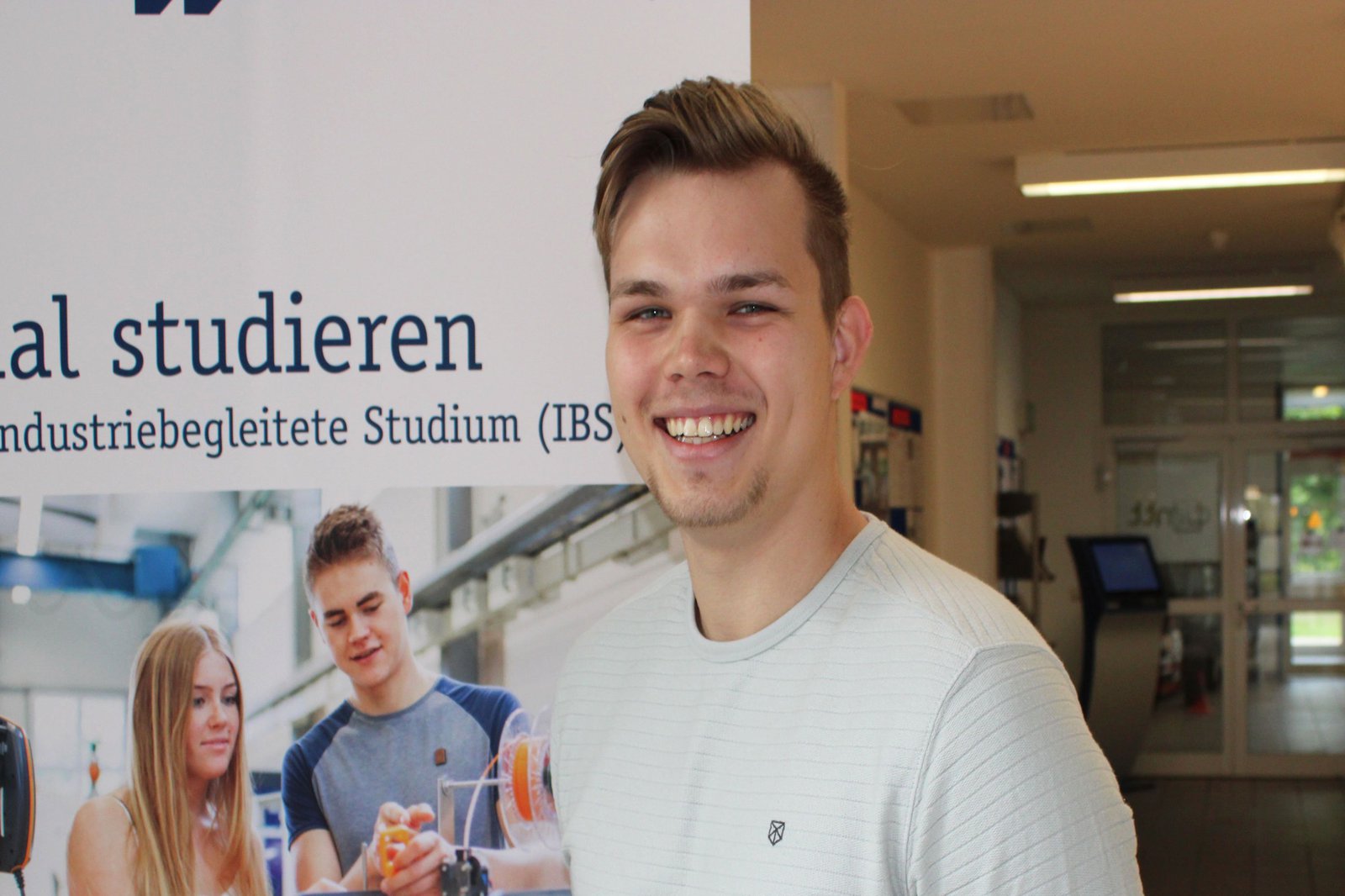 Ein Mann in einem blauen Pullover posiert vor einem Plakat in der FH Kiel.