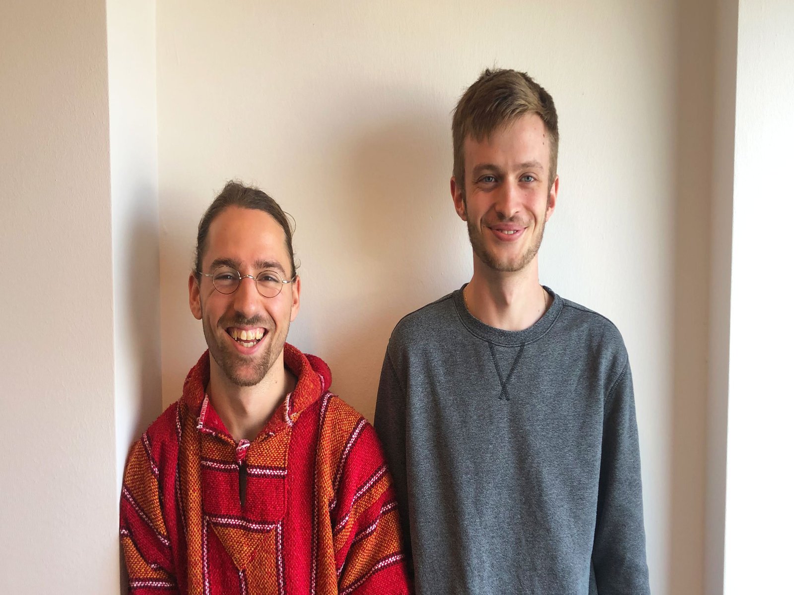 Zwei Männer posieren lächelnd vor einer weissen Wand. Der linke trägt einen roten, der rechte einen grauen Pullover.