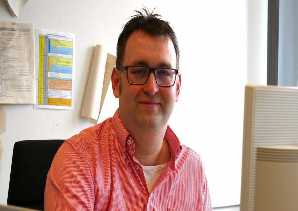 Ein Mann in rotem Hemd sitzt an seinem Arbeitsplatz und schaut in die Kamera.