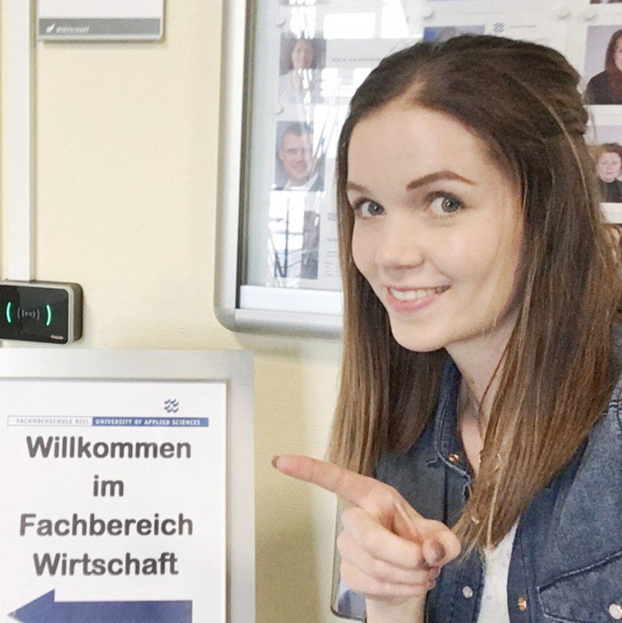 Eine Studentin zeigt mit dem Finger auf das Willkommensschild des Fachbereichs Wirtschaft der FH Kiel.