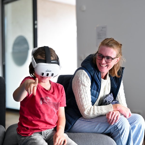 Ein Kind spielt mit einer VR-Brille. Seine Mutter schaut lachend zu.