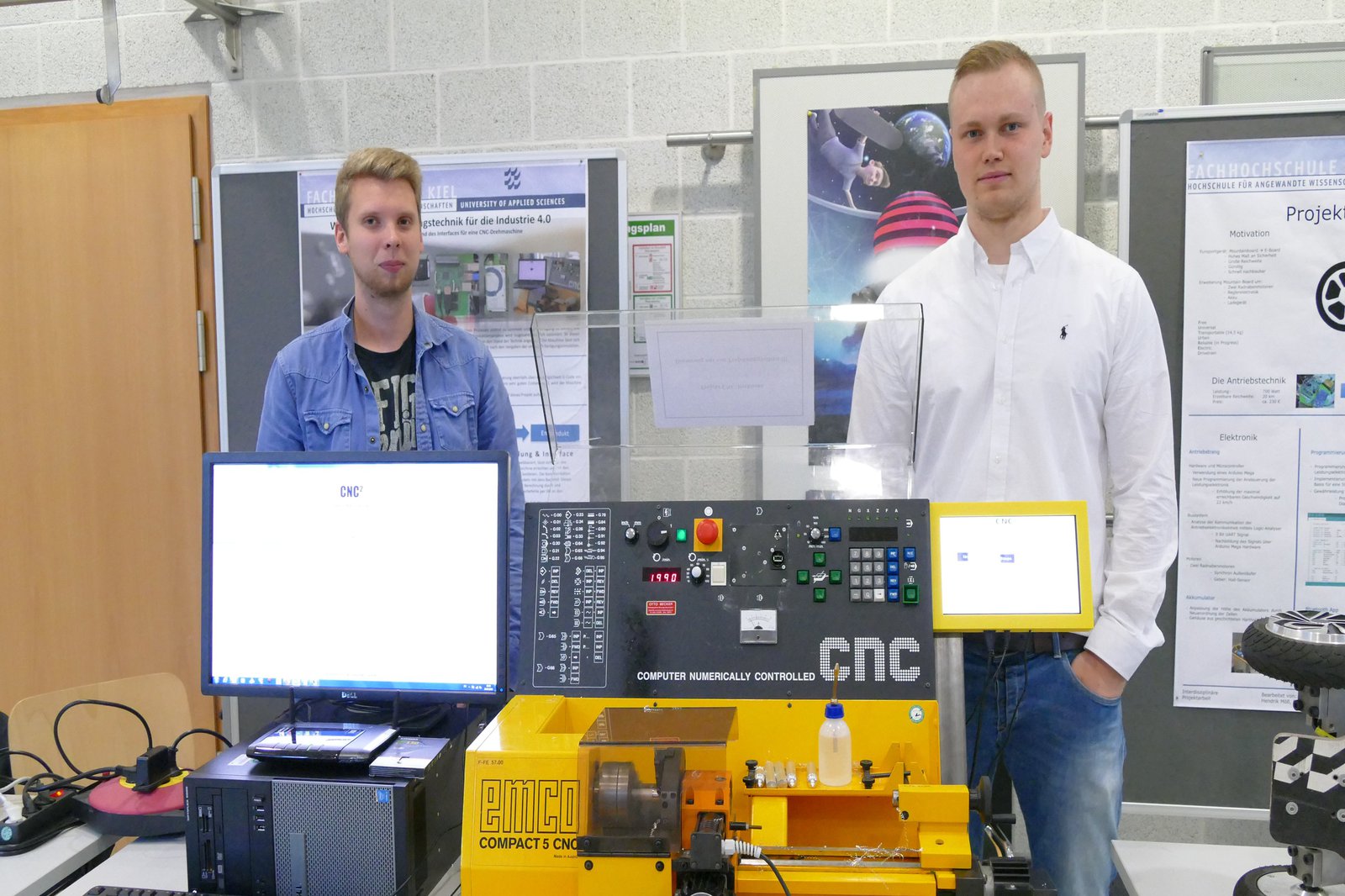 Zwei Studenten stehen hinter einer Maschine auf der "CNC" steht und einem Computer Setup.