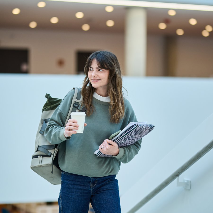 eine junge Frau mit Kaffeebecher, Schreibblock und Rucksack an der HAW Kiel