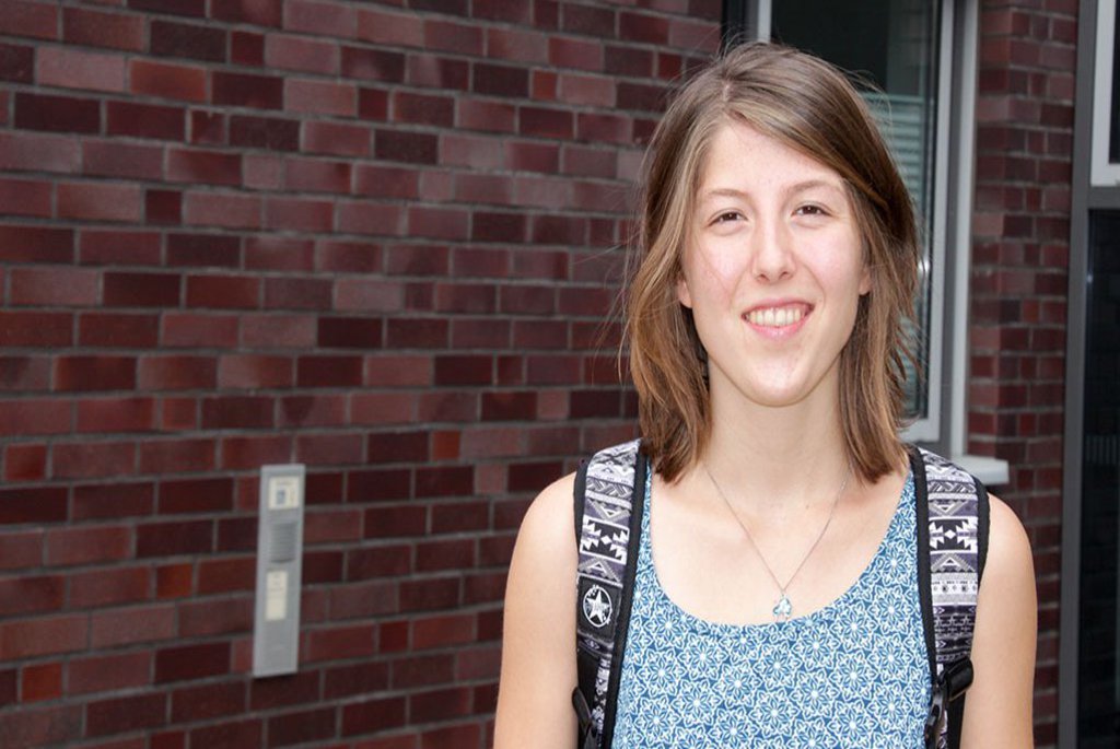 Eine Frau posiert vor einer Backsteinwand und lächelt in die Kamera.