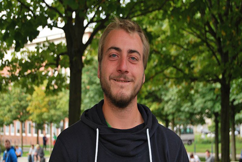 Ein Mann steht unter einem Dach aus Baumkronen und schaut zufrieden in die Kamera.