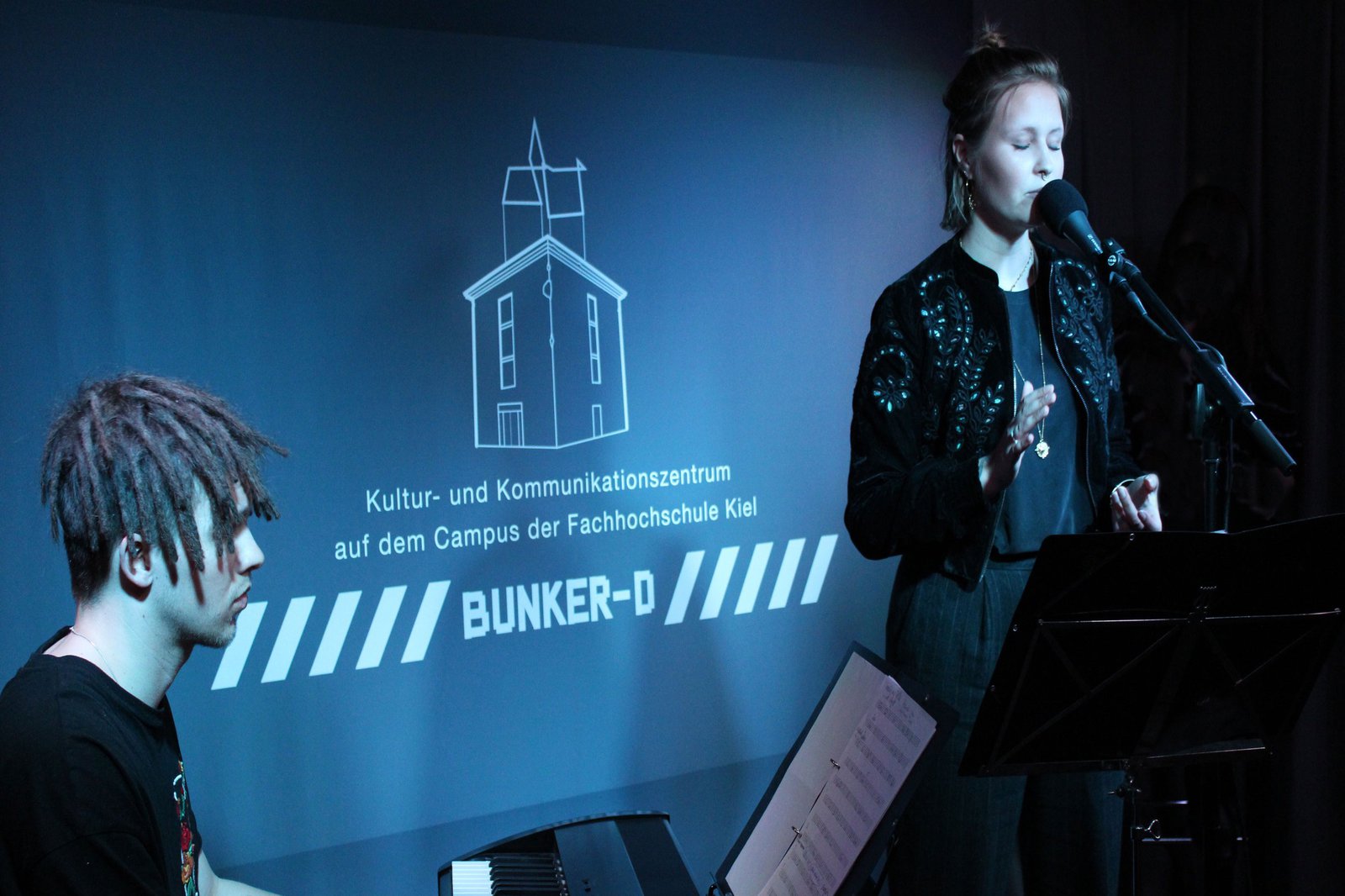 Eine Frau steht auf einer Bühne, vor einem Mikrofon, neben ihr sitzt ein Mann vor einem Klavier.