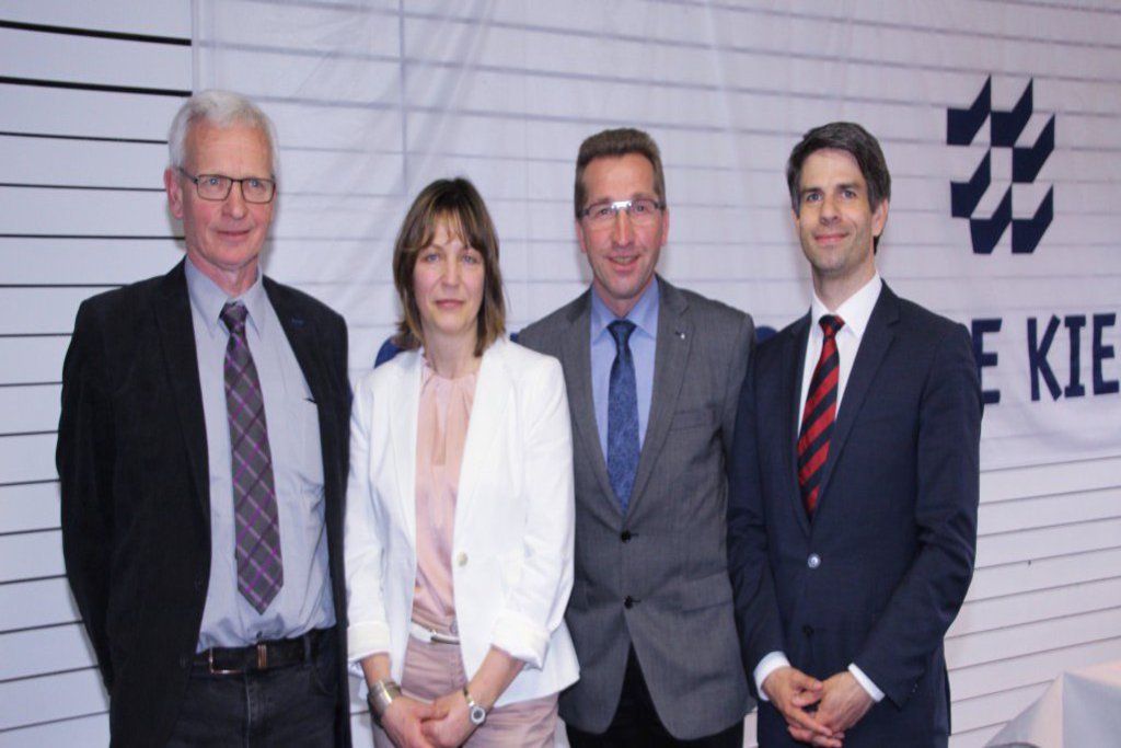 Drei Männer und eine Frau posieren vor einem Banner der "Fachhochschule Kiel".