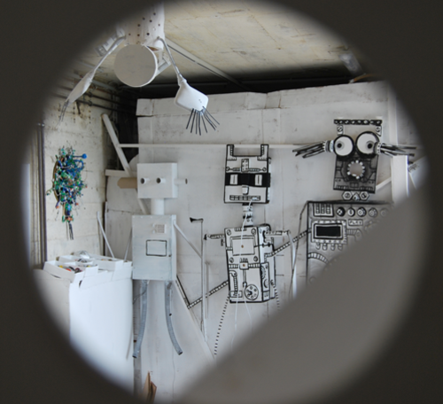 Durch ein Loch lassen sich Papp-Roboter an einer weißen Wand sehen.