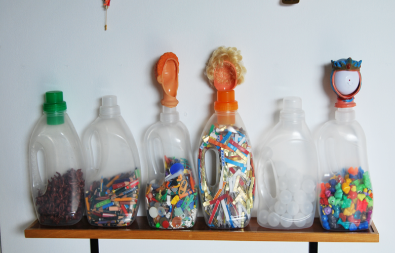 Auf einem kurzen Regal stehen sechs Waschmittelgefäße, die mit Spielzeug befüllt sind.