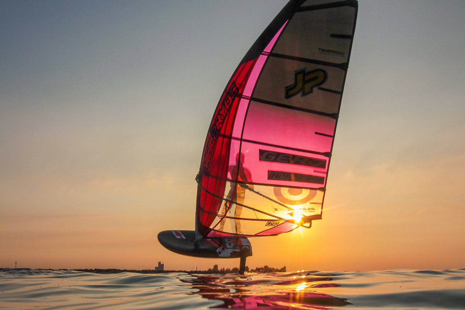 Ein über der Wasseroberfläche schwebender Windsurfer, gleitet durch die untergehende Sonne.