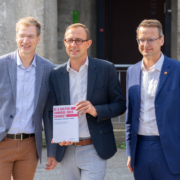 Drei Männer stehen vor dem Computermuseum der FH Kiel. Prof. Martin Lätzel (links) und Prof. Tobias Hochscherf (rechts). In der Mitte steht Digitalisierungsminister Dirk Schrödter und hält ein Buch in der Hand.