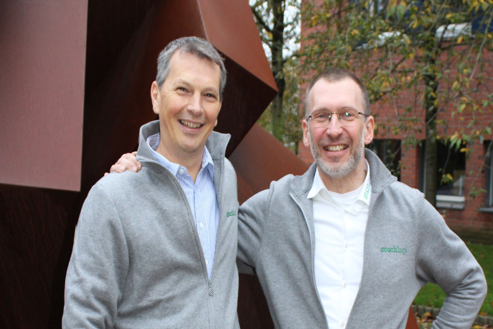 Zwei Männer in grauen Trainingsjacken, auf denen Coach steht