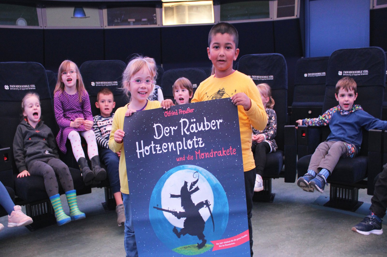 Zwei Kinder halten das Plakat zur Vorstellung "Räuber Hotzenplotz" in den Händen. Sie stehen in Mediendom und hinter ihnen befinden sich ihre Gruppenkameraden.