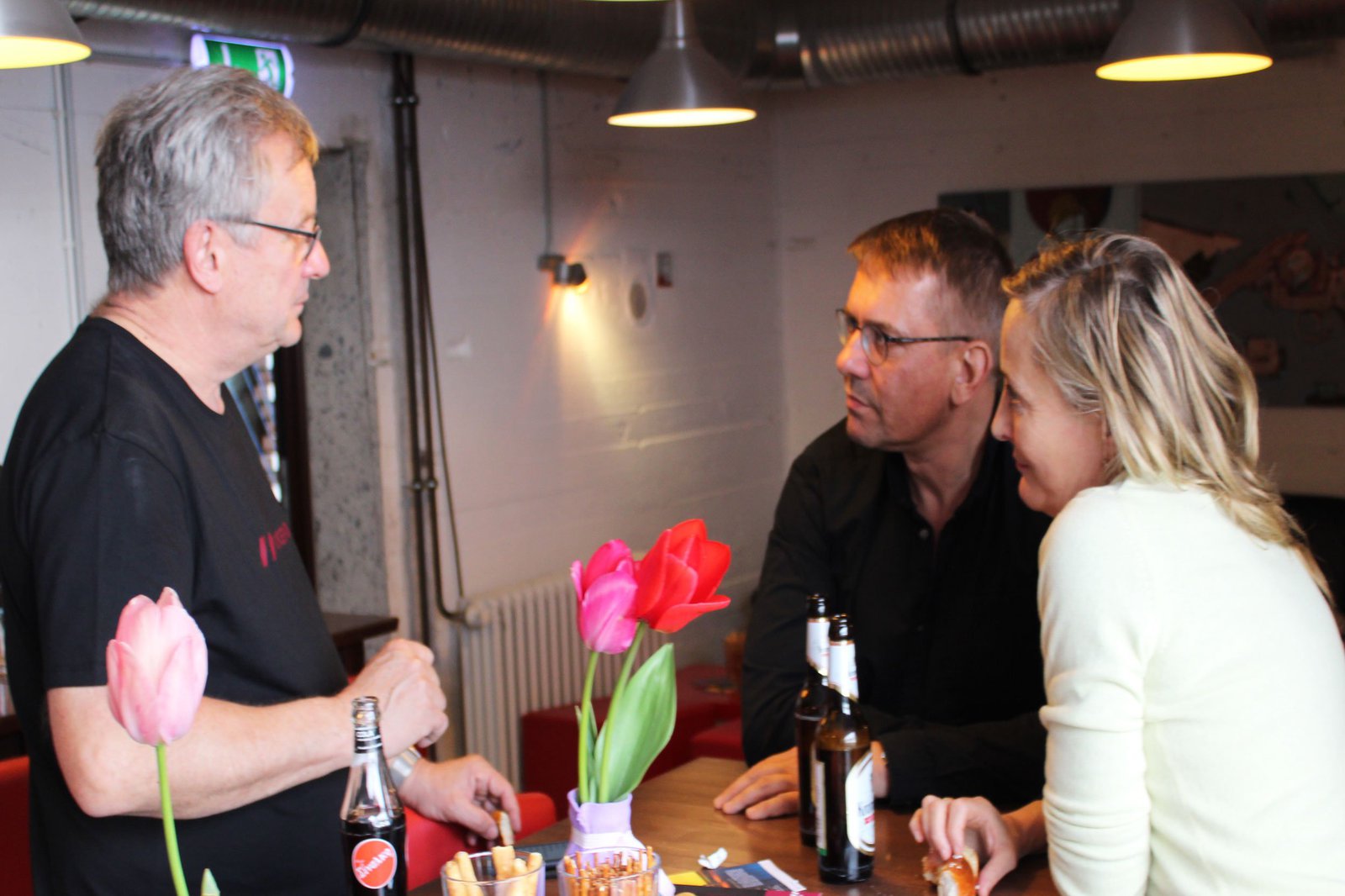 Eine Frau und zwei Männer stehen an einem Tisch, mit Getränken und Salzgebäck.