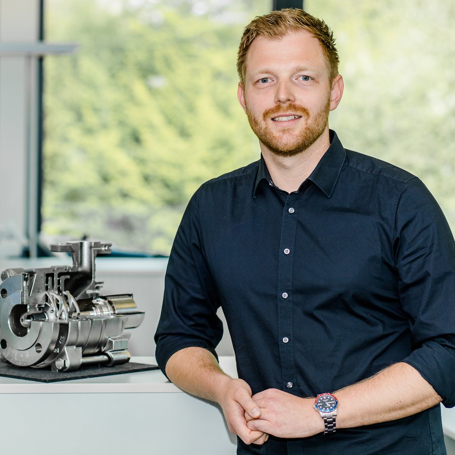 Ein junger Mann lehnt an einem Tisch und schaut lächelnd in die Kamera, auf dem Tisch steht ein Teil einer industriellen Pumpe.
