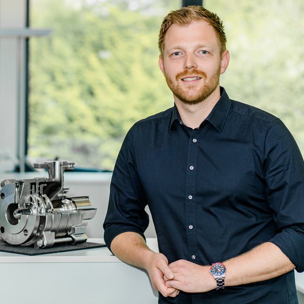 Ein junger Mann lehnt an einem Tisch und schaut lächelnd in die Kamera, auf dem Tisch steht ein Teil einer industriellen Pumpe.