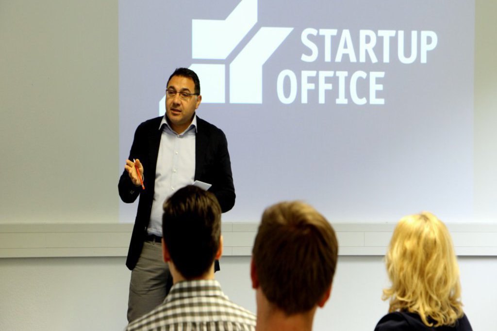 Ein Mann spricht auf einer Bühne zu dem Publikum. Der Beamer wirft das Logo des Startup Office an die Wand.