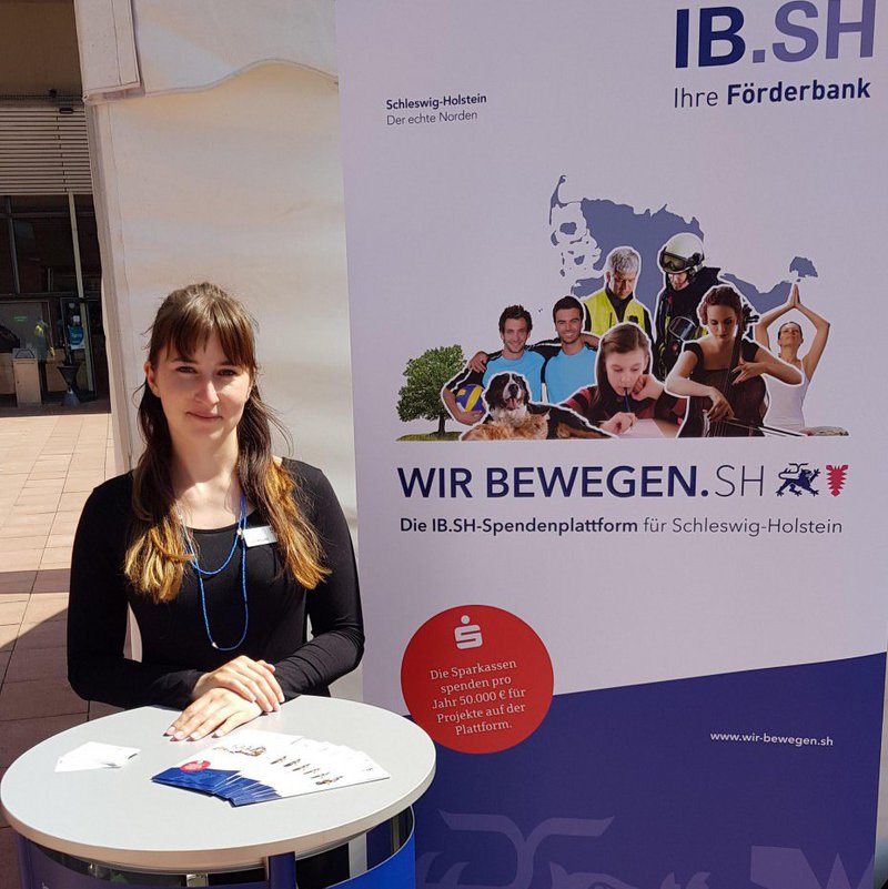 Eine Frau steht an einem Infotisch, neben ihr steht ein Plakat.