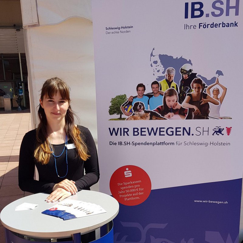 Eine Frau steht an einem Infotisch, neben ihr steht ein Plakat.