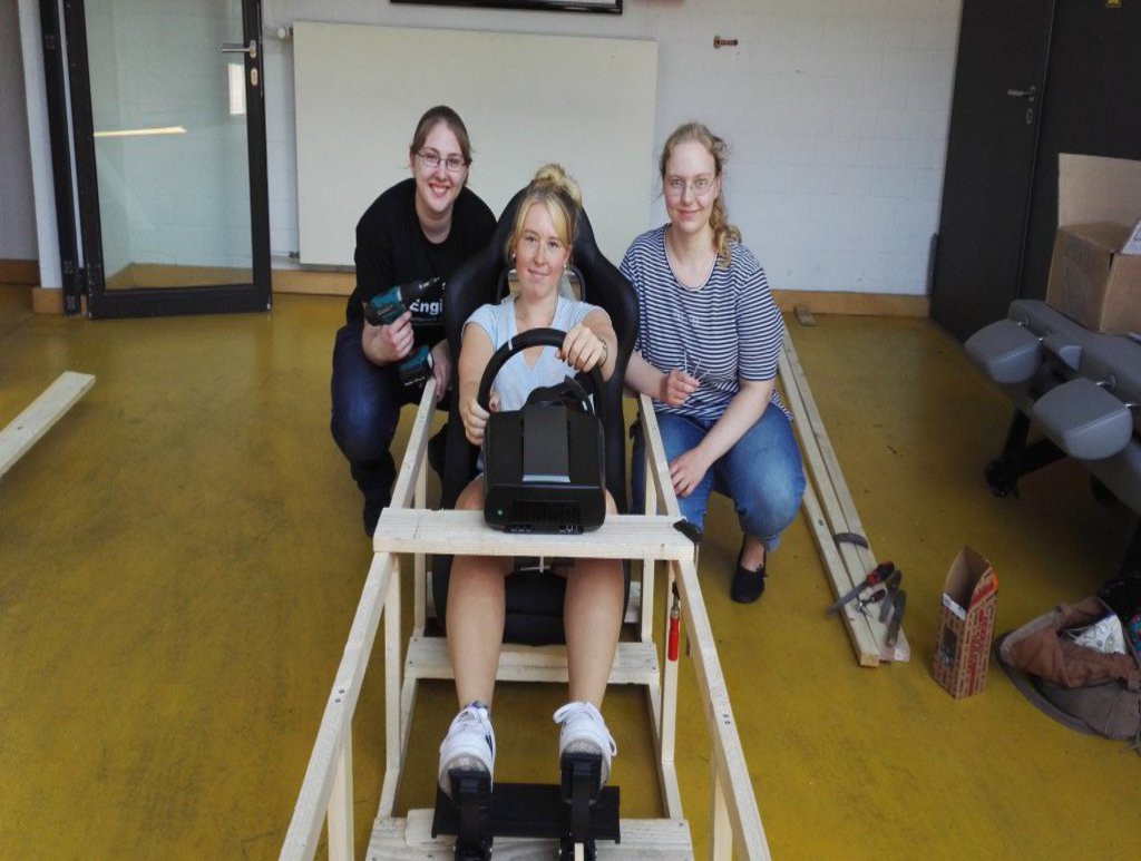 Eine Frau sitzt, von zwei Komillitoninnen umgeben, in einer nach einem Fahrzeug aussehenden Holzkonstruktion.