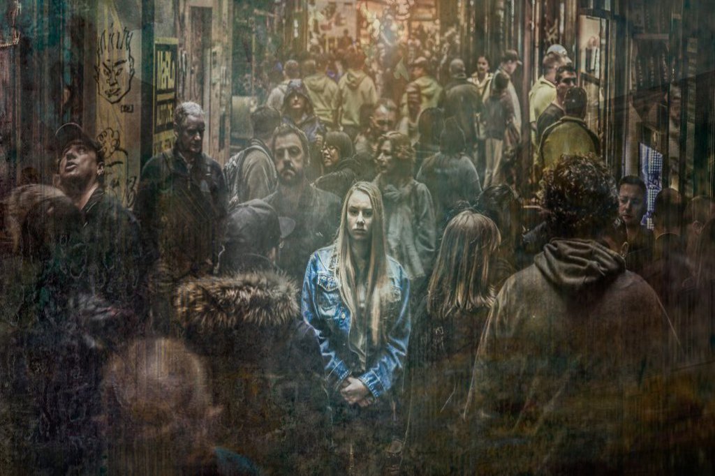 Eine FRau steht mit verschränkten Armen in einer Gasse wo sie von Menschen umgeben ist.