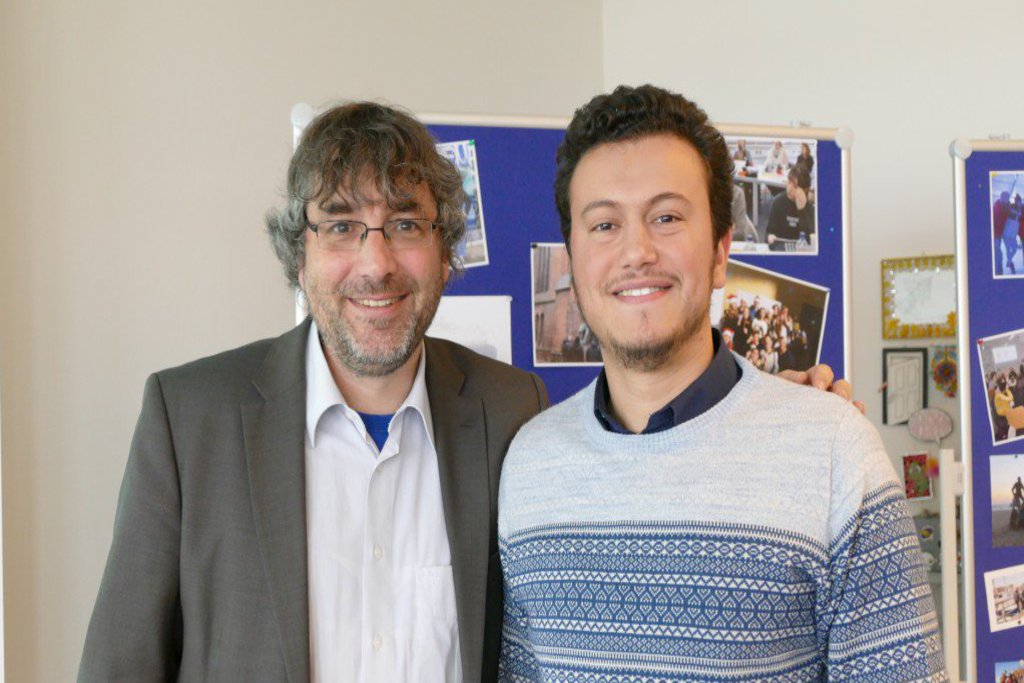 Ein älterer und ein jüngerer Mann posieren vor einer Infowand.