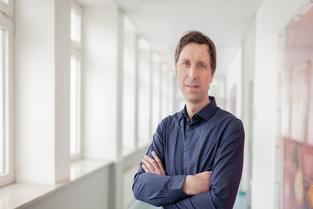 Ein Mann mit verschränkten Armen und einem blauen Hemd posiert in einem Flur der FH Kiel.