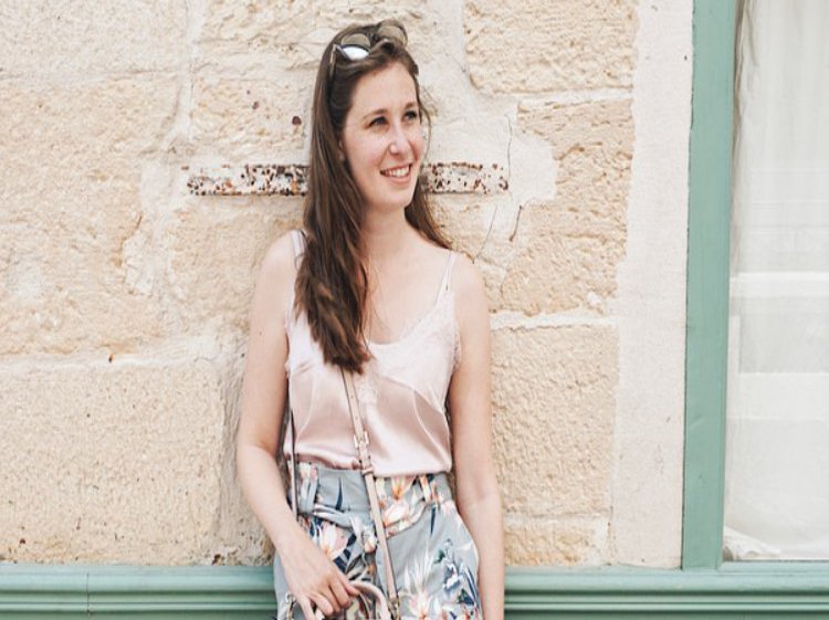 Eine Frau in einem hellen Outfit posiert an einer türkis- und aprikofarbenen Wand.