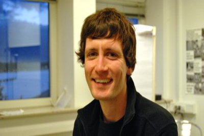 Ein Mann in blauem Pullover sitzt in einem Labor. Auf seinem Pullover lässt sich die Französische Flagge erahnen.