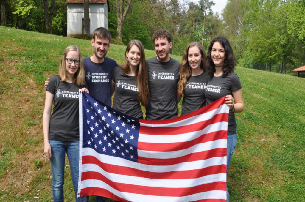 Eine Gruppe Studierender, in passenden T-Shirts, hält eine USA Flagge und posiert für die Kamera.