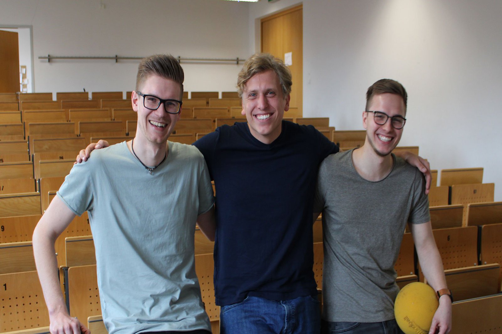 Drei Männer posieren lächelnd in einem leeren Hörsaal.