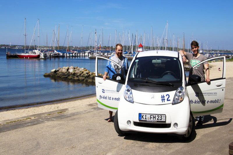 Zwei Männer stehen bei geöffneten Türen an einem E-Auto. Im Hintergrund ist ein Strandabschnitt zu sehen.