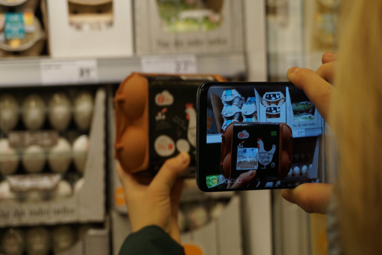 eine Frau hälft einen Eierkarton in der Hand, den sie in die Kamera ihres Handys hält; im Display ist das Video aus dem dazugehörigen Hühnerstall zu sehen