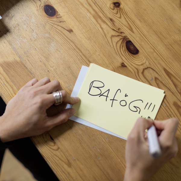 JEE_151009_DSW-Berlin_0684 Blick auf einen Tisch mit einem gelben Post-it, auf den eine Frau gerade das Wort BAföG geschrieben hat. Ihre Hände Hände rahmen den Zettel ein.