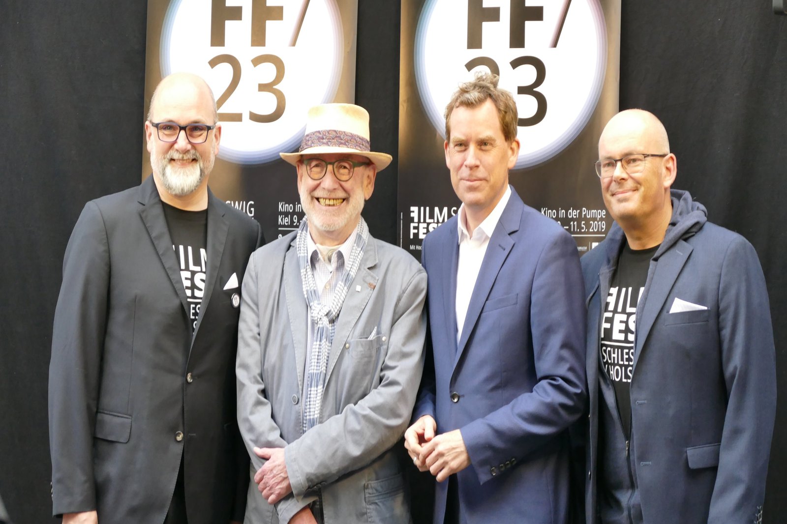 Vier Männer in Jackets posieren vor zwei Plakaten.