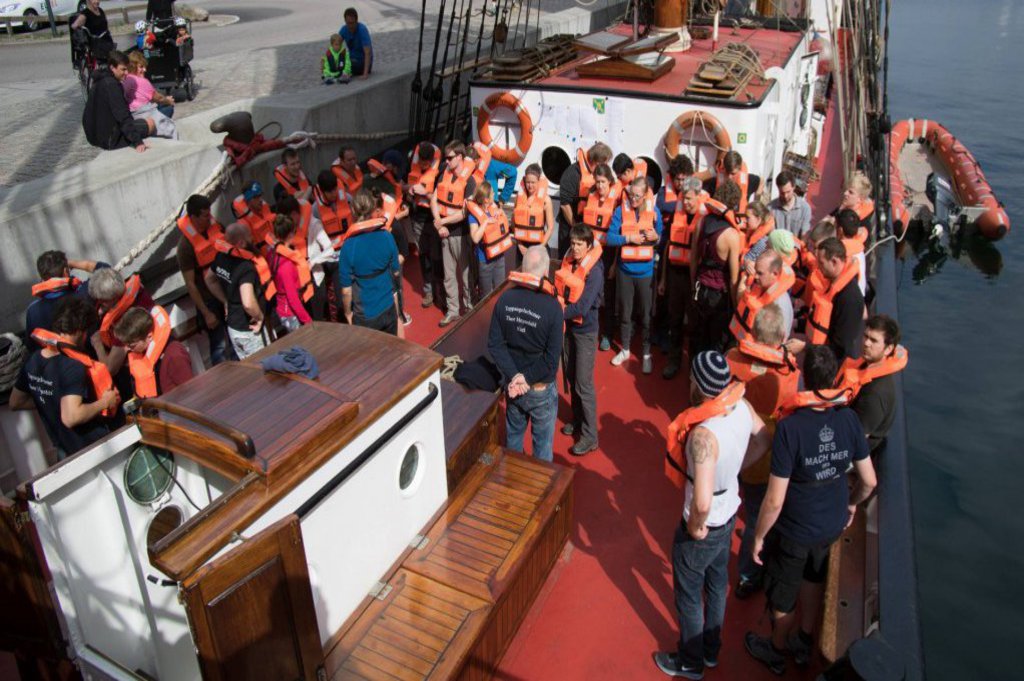 Eine Gruppe Studierender befindet sich auf einem Segelschiff. Alle sind mit einer orangenen Rettungsweste ausgestattet.
