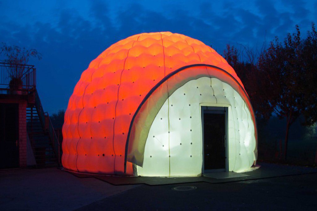 Ein großes Zelt in der Form eines Iglu, ist mit einer orangenen Plane überdeckt und von innen stark beleuchtet.