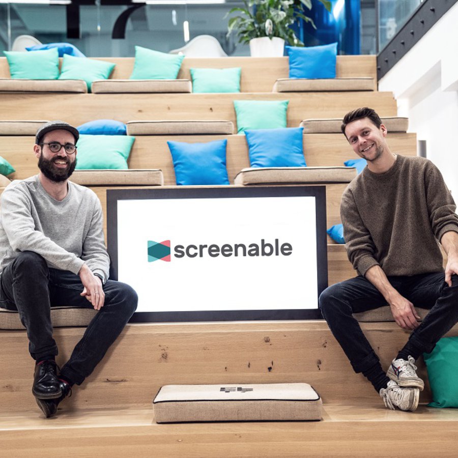 Jens Woltering und Philipp Spieck sitzen auf einer Treppe, zwischen ihnen steht der Prototyp ihres interaktiven Screens