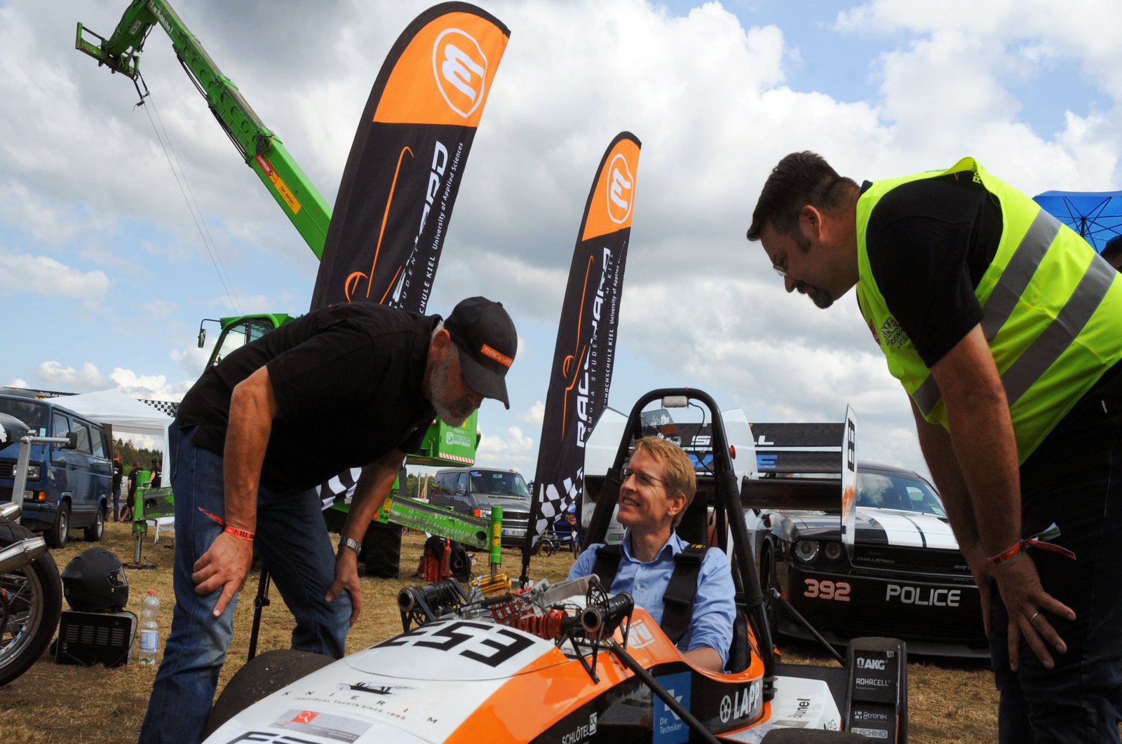 Daniel Günther sitzt freudig in einem Rennwagen und lässt sich von zwei Männern informieren.