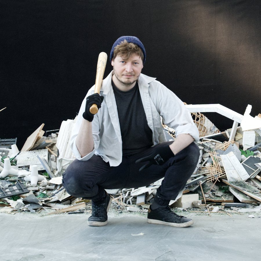 Ein junger Mann mit Basecap hockt, einen Baseballschläger in der Hand, vor einem Haufen zerschlagener Gegenstände