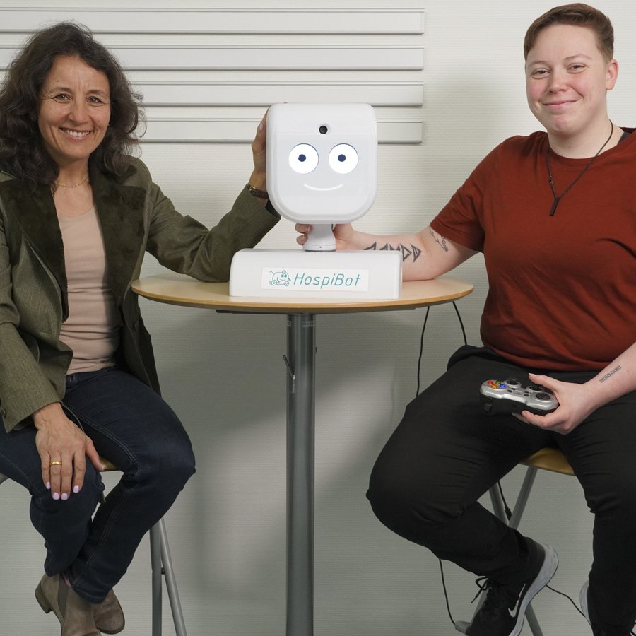 Zwei Frauen sitzen auf der rechten und linken Seite eines Bistrotischs, auf dem Tisch steht der Kopf eines Roboters.