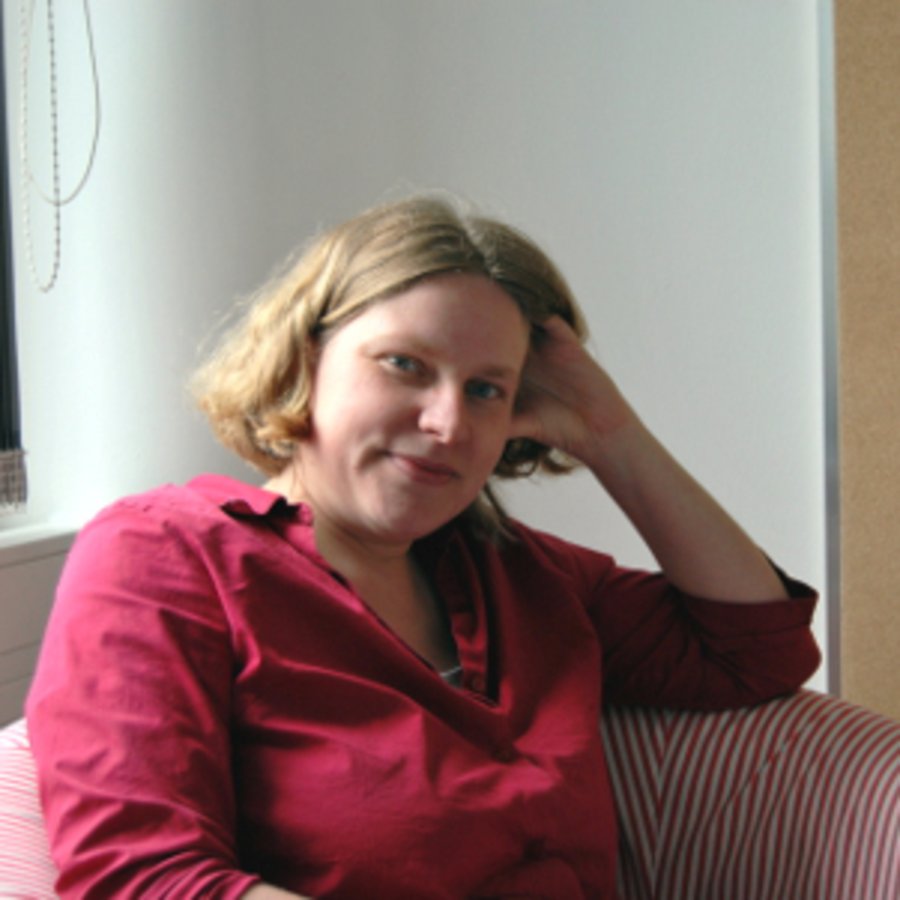 Eine Frau mit rotem Hemd und blondem Bob, posiert auf einem Sessel für die Kamera.