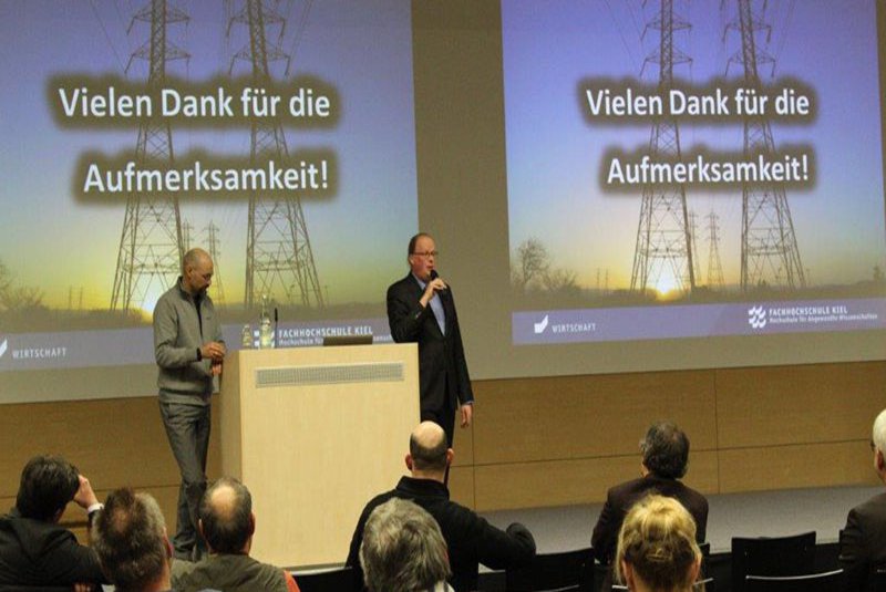 Zwei Männer stehen auf einer Bühne. Einer von ihnen hält ein Mikrofon in den Händen und spricht zum Publikum.