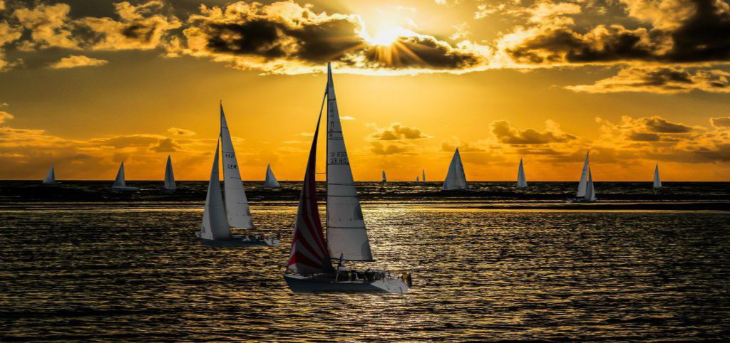 Eine vielzahl an Segelbooten schippernd vor einem goldenen Sonnenuntergang.