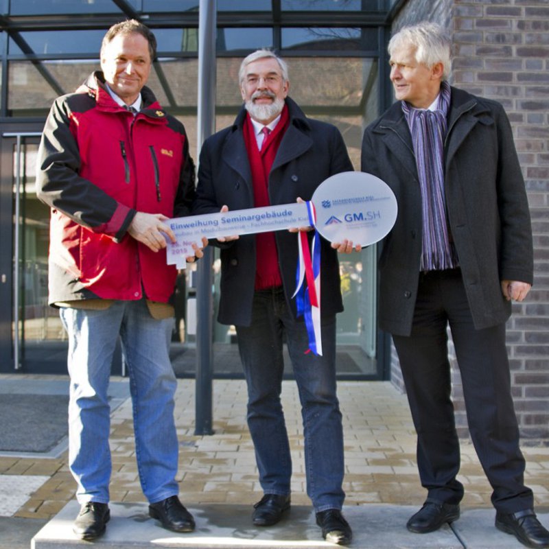 Drei ältere Männer in Winterkleidung, halten einen überdimensionierten Schlüssel in den Händen.